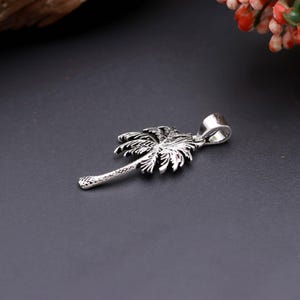 Könnte beinhalten: Ein silberfarbener Metallanhänger in Form einer Palme. Das Design zeigt einen strukturierten Stamm und detaillierte Wedel an den Blättern. Eine glatte, polierte Metallöse ist an der Oberseite der Palme zur Befestigung an einer Kette angebracht.