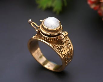 May include: A gold-toned metal ring featuring an intricate floral pattern engraved along the band. The top of the ring showcases a circular, hinged locket-style compartment topped with a smooth, round, white iridescent gemstone cabochon.