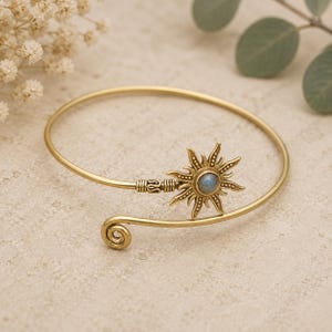 Puede incluir: Un brazalete dorado con un diseño de sol. El sol presenta una piedra central redonda de color azul claro. El brazalete tiene un diseño en espiral en un extremo y un cierre decorativo en el otro.