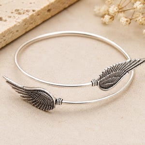 Könnte beinhalten: Silberfarbenes Armband mit zwei detaillierten Flügelmotiven. Die Flügel sind auf beiden Seiten des offenen Armreifs angeordnet und bilden ein symmetrisches Design. Das Armband ist vor einem neutralen Hintergrund platziert, was seine filigrane Verarbeitung hervorhebt.