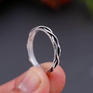 Peut inclure: Une bague argentée présentant un motif torsadé rappelant une corde, avec des zones sombres et creuses entre les brins. L'anneau en métal possède une finition texturée et oxydée qui souligne le motif entrelacé sur toute sa circonférence.