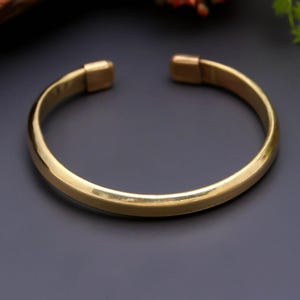 Può includere: Un bracciale rigido in metallo color oro lucido con una superficie liscia e arrotondata e un design aperto. Il bracciale presenta due terminali arrotondati leggermente più spessi all'apertura, creando una struttura circolare continua.