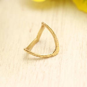 Pode incluir: Um anel de tom dourado com um design em forma de V e uma superfície martelada e texturizada. A banda é aberta no topo, formando uma ponta em chevron. O metal tem um acabamento reflexivo que capta a luz, destacando a textura irregular e com covinhas em toda a superfície externa da banda do anel.