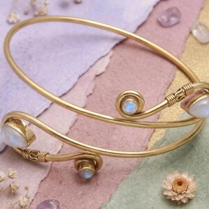 May include: A gold-colored cuff bracelet with three moonstone accents. The bracelet has a unique, open design with curved lines and decorative detailing. The moonstones are pearlescent and add a touch of elegance to the piece. The bracelet is displayed on a colorful, textured surface.