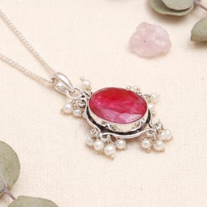 Op de afbeelding: Een zilveren hangerketting met een grote, ovaal geslepen robijn edelsteen. De robijn is omgeven door kleine, witte parels en gezet in een decoratief zilveren frame. De ketting wordt getoond tegen een neutrale achtergrond met groene takjes.