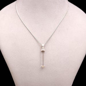 May include: A clear quartz crystal pendant necklace. The pendant is a clear, elongated crystal with a silver-coloured cap, suspended from a delicate silver chain. The necklace is displayed on a white mannequin.