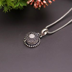 May include: Silver-toned pendant necklace featuring a round, locket-style charm. The charm has a floral design around a central, light-colored stone. The necklace chain is a delicate, linked style. The background is a dark gray.