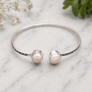 May include: A silver open cuff bracelet with a pearl at each end. The bracelet has a textured band and is set against a marble background. The pearls are a creamy white colour.