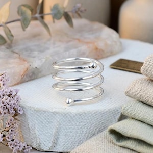 Könnte beinhalten: Ein silberner Spiralring mit einer kleinen Kugel an jedem Ende. Der Ring besteht aus glänzendem Metall und hat ein gewendeltes Design. Der Ring wird auf einer weißen Oberfläche mit verschwommenem Hintergrund präsentiert.