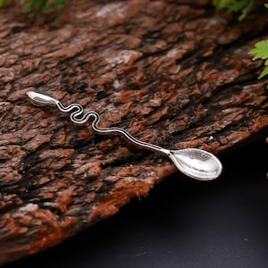 Puede incluir: Una cuchara de metal plateado con un mango largo y delgado que presenta un diseño ondulado y serpenteante. El mango termina en una pequeña punta redondeada en un extremo y en un cuenco ovalado poco profundo en el otro, con acabado metálico.