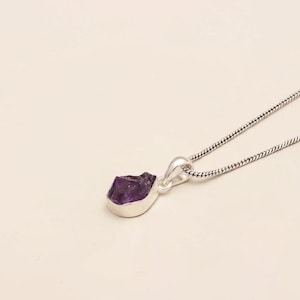 May include: A silver necklace featuring a raw, deep purple amethyst gemstone pendant set in a smooth silver bezel. The pendant is attached to a thin, flexible silver snake chain that extends diagonally across the frame from the upper right corner.