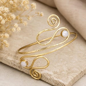 May include: Gold-coloured arm cuff with a swirling design and two small, round, white gemstones. The cuff features hammered metal detailing and a spiral accent. A stylish piece of jewellery.