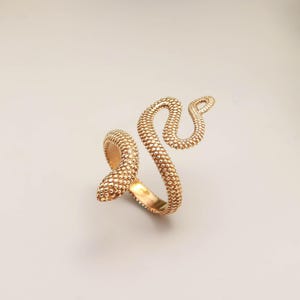 Puede incluir: Un anillo de serpiente dorado con un patrón texturizado en forma de escamas. El anillo se enrolla alrededor del dedo en un diseño de serpiente estilizado, con la cabeza y la cola curvadas hacia arriba. Es una joya.