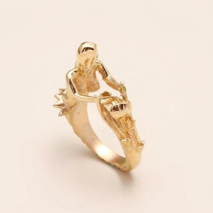 May include: A gold-colored ring featuring a mermaid design. The ring depicts a mermaid holding a basket, with intricate details on the mermaid and the ring's band. The ring is made of a shiny, metallic material.