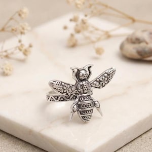May include: A silver-toned metal ring featuring a detailed bee design on the front. The bee has textured wings with floral patterns and a striped abdomen. The ring band displays a geometric triangle pattern along its visible outer surface.