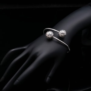 Op de afbeelding: Een zilverkleurige open armband met twee bolvormige uiteinden. Elke bol is voorzien van een geometrisch uitgesneden patroon dat doet denken aan een levensbloem. De armband heeft een gladde, dunne band die rond de pols buigt.
