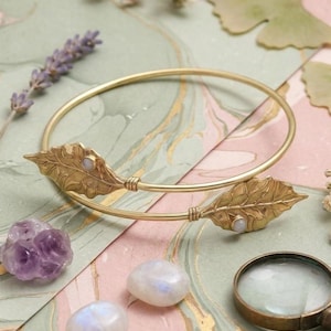 Op de afbeelding: Een goudkleurige armband met bladvormige accenten en kleine, ronde edelstenen. De armband wordt getoond met amethist, maanstenen en een vergrootglas op een patroonoppervlak.