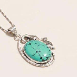 Pode incluir: Um colar com pendente de prata apresentando uma grande pedra turquesa oval com veios pretos. A pedra está engastada numa moldura de prata decorada com uma pequena figura de pássaro e um motivo de serpente, presa a uma fina corrente de prata.