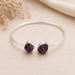 Könnte beinhalten: Ein silberfarbenes Armreif-Armband mit zwei rohen, dunkelvioletten Amethyststeinen. Das Armband hat ein schlichtes, offenes Design mit glatter, polierter Oberfläche. Die Amethyststeine sind in Silberfassungen gefasst.