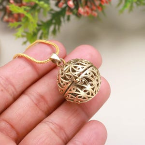 以下が含まれることがあります： 金色のペンダントネックレス。透かし細工の球体デザインが特徴です。球体は精巧なオープンワーク模様で、繊細なゴールドチェーンから吊り下げられています。柔らかい背景を背景にネックレスが展示されており、ジュエリーの職人技が際立っています。