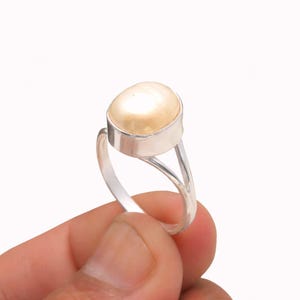 Könnte beinhalten: Ein Silberring mit einem großen, ovalen, cremefarbenen Edelstein. Der Edelstein ist in einer silbernen Lünette gefasst und der Ring hat ein geteiltes Schaftdesign. Der Ring wird in einer Hand vor weißem Hintergrund gehalten.