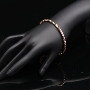 Op de afbeelding: Een metalen armband met een gedraaid touwontwerp met afwisselende banden in koper-, zilver- en goudtinten. De cirkelvormige armband heeft een gepolijst, reflecterend oppervlak dat het licht opvangt en een veelkleurig uiterlijk creëert.