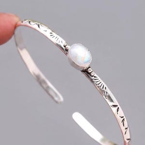 Könnte beinhalten: Ein silbernes Armreif-Armband mit einem ovalen, irisierenden Edelstein in der Mitte. Das Armband hat geätzte dekorative Muster entlang des Bandes. Das offene Design ermöglicht ein einfaches Tragen. Ein schlichtes und elegantes Schmuckstück.