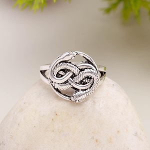 May include: A silver ring with a detailed design of two intertwined snakes in a circular form. The snakes have textured scales and are arranged symmetrically. The ring is displayed on a light-coloured, textured surface.
