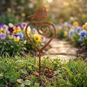 Könnte beinhalten: Ein rostiger Metall-Gartenstecker mit einem Vogel, der auf einem herzförmigen Design mit einer Spirale sitzt. Der Stecker befindet sich in einem Garten mit bunten Blumen und Gras und dient als dekoratives Element im Freien.
