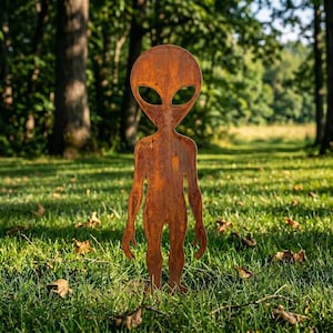 Könnte beinhalten: Eine flache, ausgeschnittene Metallskulptur einer Alien-Figur mit einem großen, ovalen Kopf und zwei hohlen, mandelförmigen Augenhöhlen. Die gesamte Oberfläche der Figur weist ein strukturiertes, verwittertes und rostbraunes Patina-Finish auf.