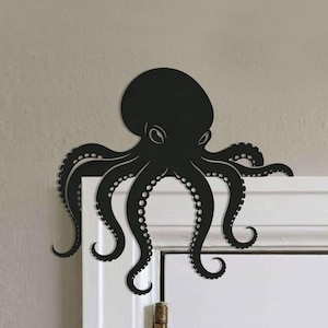 Op de afbeelding: Een matzwart metalen wanddecoratie in de vorm van een octopus, ontworpen om op de hoek van een deurkozijn te worden gemonteerd. De octopus heeft een grote ronde kop, twee zichtbare ogen en acht gebogen tentakels met ronde zuignapdetails.