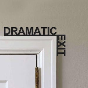 May include: A black, cut-out wall sign featuring the text "DRAMATIC EXIT" in a bold, sans-serif font. The word "DRAMATIC" is positioned horizontally above the door frame, while the word "EXIT" extends vertically down the side of the frame.
