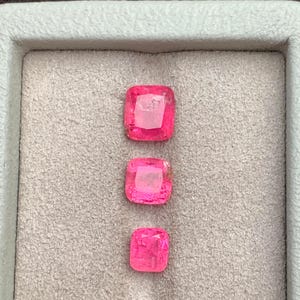 Op de afbeelding: Drie levendige, vierkant geslepen, felroze edelstenen worden getoond in een lichtgrijze tray. De edelstenen zijn verticaal gerangschikt, elk met een iets andere grootte en interne facetten, wat hun schittering laat zien. De tray staat tegen een neutrale achtergrond.