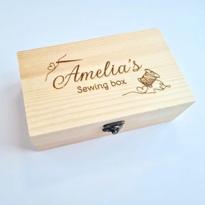 May include: A rectangular light-coloured wooden sewing box with a hinged lid and a dark metal latch. The lid features laser-engraved text that reads Amelia's Sewing box, accompanied by illustrations of a needle, thread spool, and small hearts.