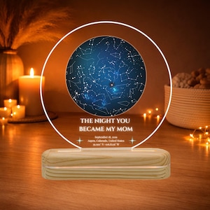Puede incluir: Un mapa estelar personalizado exhibido en un panel acrílico redondo e iluminado. El texto dice "THE NIGHT YOU BECAME MY MOM" con una fecha y ubicación. La base es de madera clara.