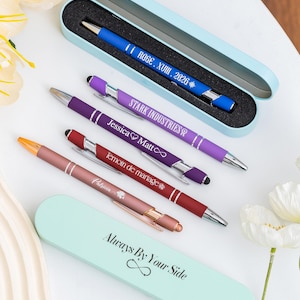Puede incluir: Un surtido de bolígrafos personalizados en varios colores, incluyendo azul, morado y oro rosa, con detalles plateados. Cada bolígrafo presenta texto personalizado, como nombres y fechas, y descansa en un estuche a juego. El estuche dice "Always By Your Side".
