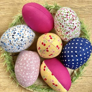 May include: A collection of seven fabric Easter eggs in various colors and patterns. The eggs include solid pink, floral prints, and polka dots, arranged on a bed of greenery. The eggs are a mix of oval and round shapes.