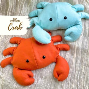 May include: Two plush crab toys, one light blue and one orange, with black button eyes. The crabs are made of soft fabric and have detailed claws and legs. The text "PDF PATTERN Crab" is on the left side of the image.