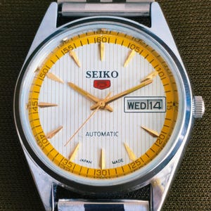 May include: A silver Seiko automatic wristwatch with a white face and gold accents. The watch face features gold hour markers, hands, and a day-date display. The watch band is silver and the watch is marked "Japan Made".