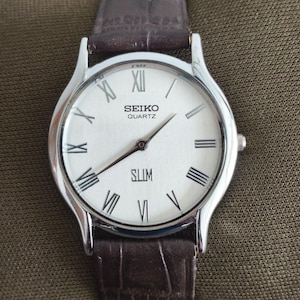 Peut inclure: Une montre Seiko Quartz argentée avec un cadran blanc, des aiguilles noires et des chiffres romains. Le bracelet est en cuir marron. Le mot "SLIM" est imprimé sur le cadran.