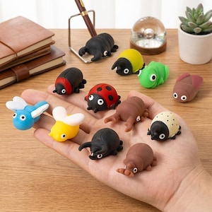 May include: A selection of colourful insect-shaped rubbers. The rubbers feature ladybirds, beetles, bees, and other bugs in various colours such as red, yellow, green, and black. The rubbers are small and detailed, with rounded shapes and simple features.