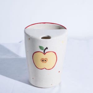 May include: A white ceramic toothbrush holder with a red rim and an apple design. The apple is yellow with a red outline and a green leaf. The holder has a hole for a toothbrush.