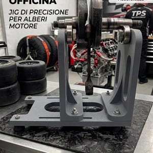 Puede incluir: Una plantilla de precisión gris para cigüeñales de motor, diseñada para la precisión. La plantilla sujeta un cigüeñal con cojinetes y pernos adjuntos. El texto en italiano dice "Mejora tu taller" y "Diseñado para la máxima precisión".