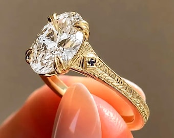 May include: A gold ring featuring a large oval-cut clear gemstone held by four prongs. The band is crafted from yellow gold with an intricate engraved leaf-like pattern and a small, square-cut dark blue gemstone set into the side of the shank.