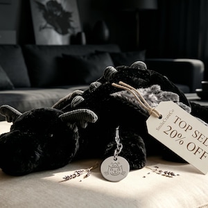 Op de afbeelding: Een zwart pluche draakspeelgoed met een label met de tekst "TOP SELL 20% OFF" en een kleine zilveren bedel. De draak ligt op een beige bank in een kamer met donkere meubels en decor.