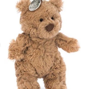 Könnte beinhalten: Ein brauner Teddybär-Schlüsselanhänger mit einem silberfarbenen Anhänger, der an einem Metallring befestigt ist. Der Bär hat ein weiches, strukturiertes Aussehen, mit einer braunen Nase und kleinen schwarzen Augen. Der Anhänger hat ein geprägtes Design.