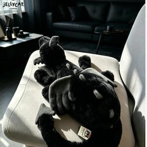 Op de afbeelding: Een zwarte pluche draak met grijze accenten, waaronder vleugels en stekels. De draak ligt op een lichtgrijze stoel. Het Jellycat-logo is zichtbaar in de linkerbovenhoek.