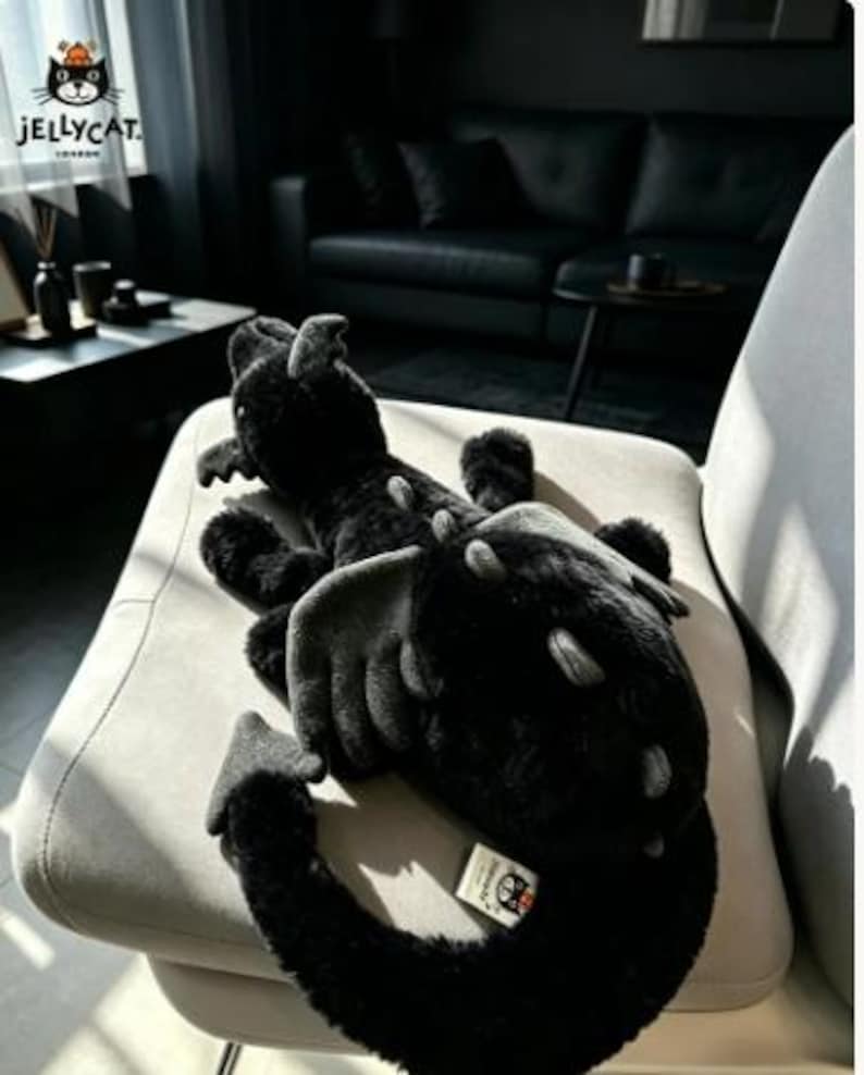Op de afbeelding: Een zwarte pluche draak met grijze accenten, waaronder vleugels en stekels. De draak ligt op een lichtgrijze stoel. Het Jellycat-logo is zichtbaar in de linkerbovenhoek.