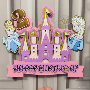 May include: A layered cake topper featuring a purple castle with gold accents, the number 2, and the words "Happy Birthday." Cinderella and Elsa are depicted on either side of the castle. The topper is held by a clear plastic stick.
