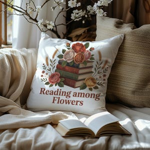 May include: White decorative pillow with a floral and book design. The pillow features a stack of books and roses in shades of red, pink, and yellow. The text "Reading among Flowers" is printed in brown. An open book rests in front of the pillow.